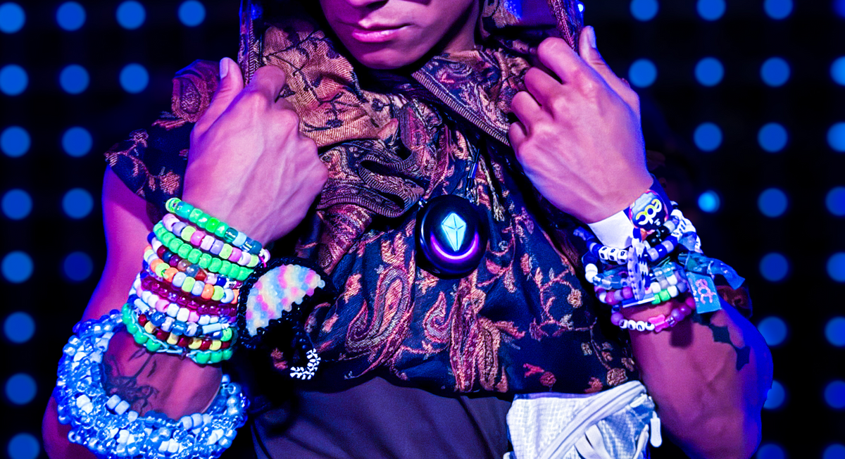 Festival-goer at a rave in a paisley scarf and stacks of kandi bracelets, wearing a Totem Compass pendant with a glowing crystal, face cropped out of frame