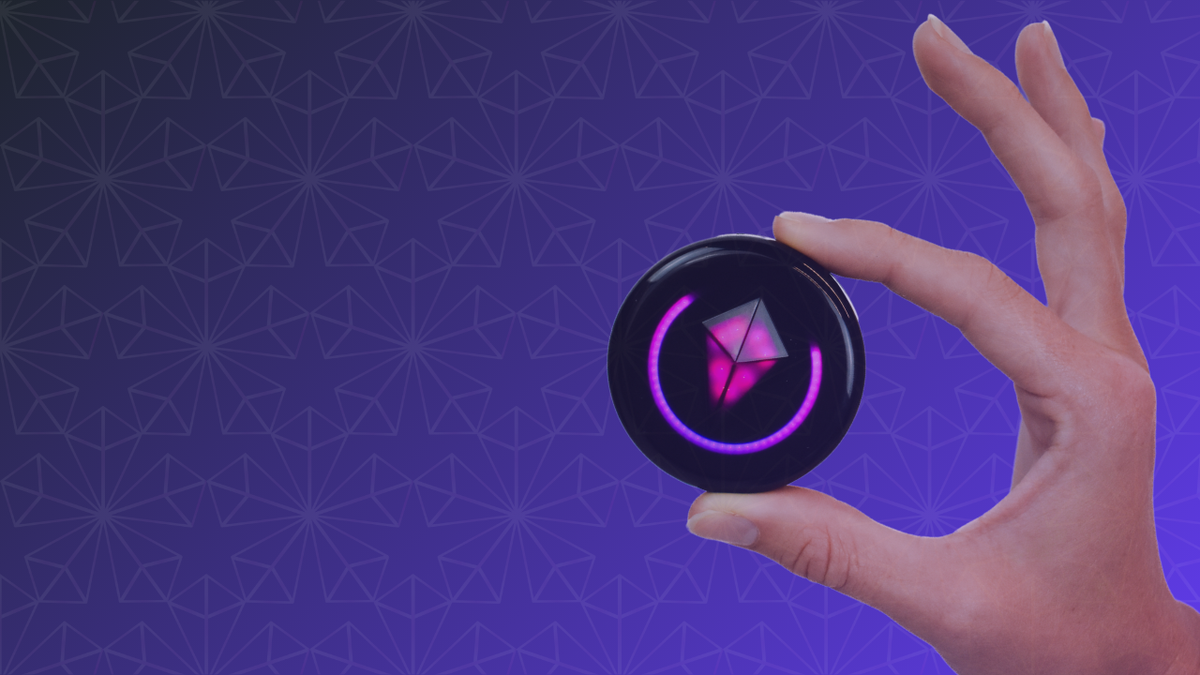 Hand holding a Totem Compass between two fingers, pink Touch Crystal and pink Halo glowing, on a purple patterned background