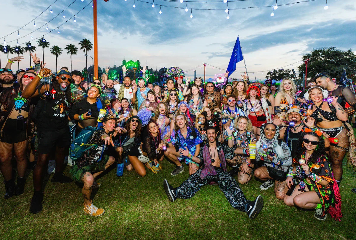 Large group of festival-goers in colorful outfits posing together, several wearing Totem Compasses