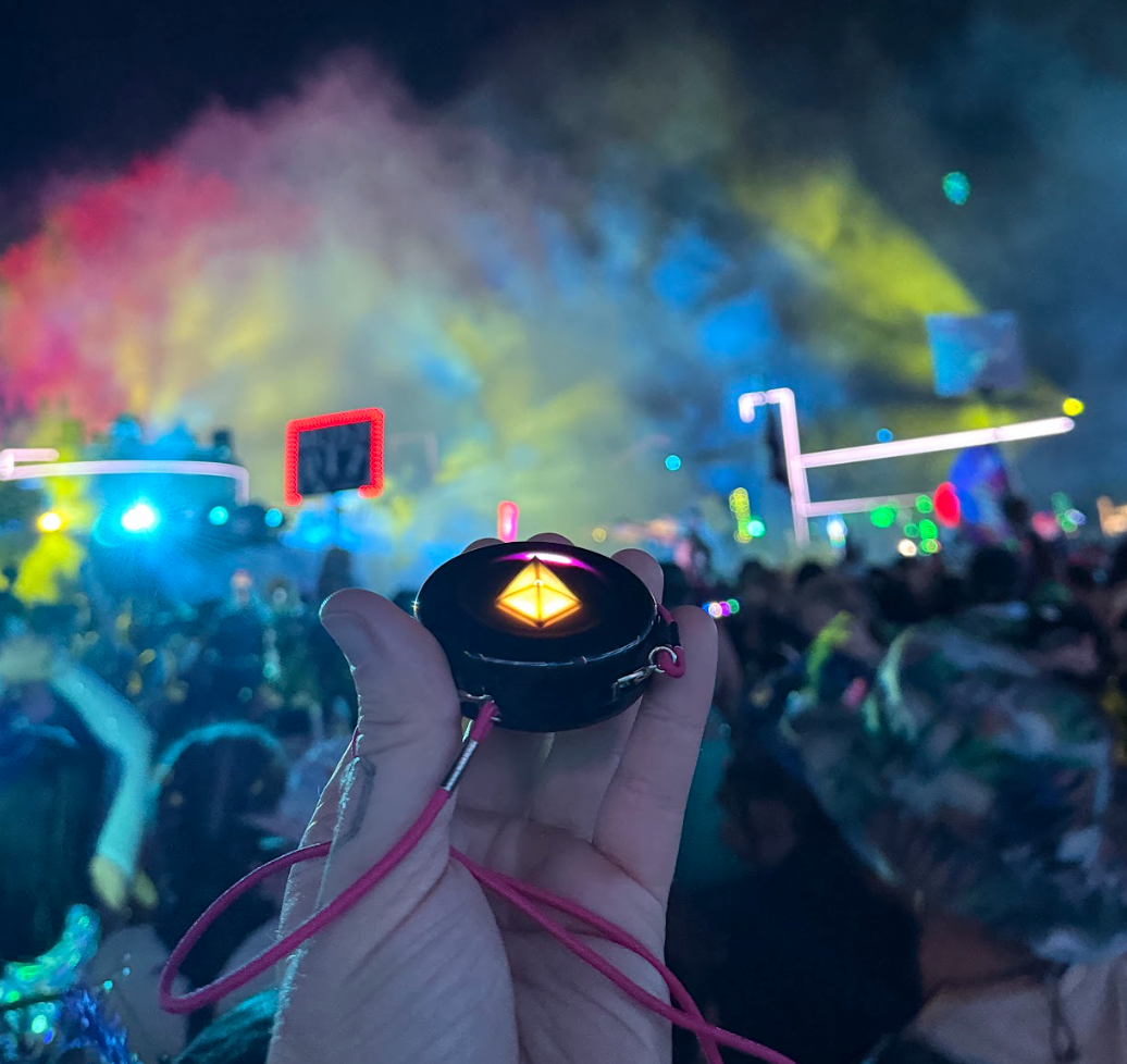 Hand holding a glowing Totem Compass on a pink cord in front of a festival stage with rainbow smoke and lights