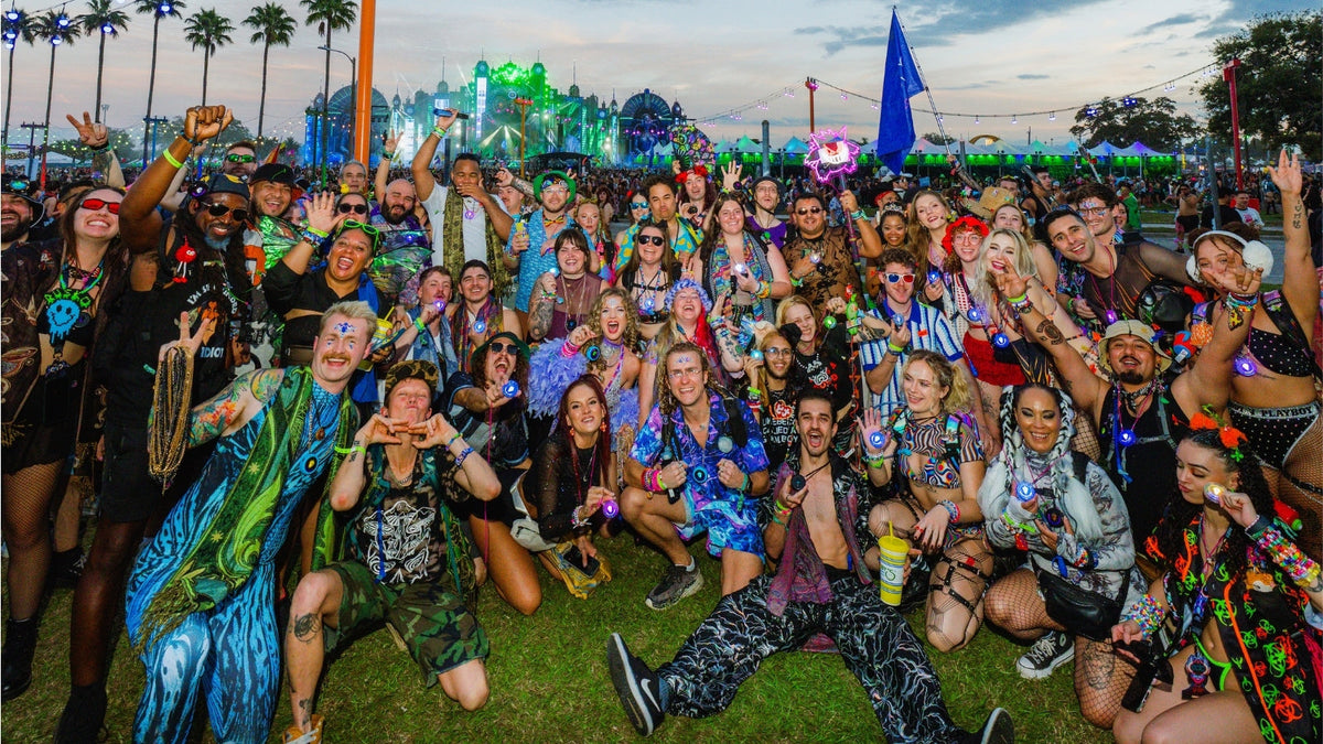Large crowd of Totem users in colorful festival outfits posing together on the grass at a Totem meetup, with a blue Totem flag and a lit-up festival skyline behind them