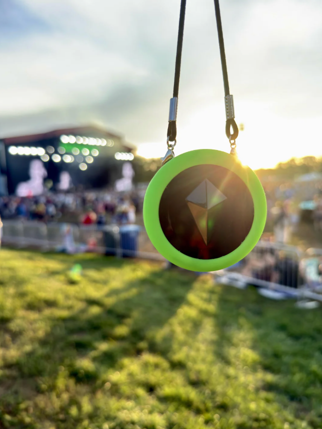 Totem Compass in a neon green Skin hanging from a black cord in the late-afternoon sun, with a blurred festival stage and crowd behind it