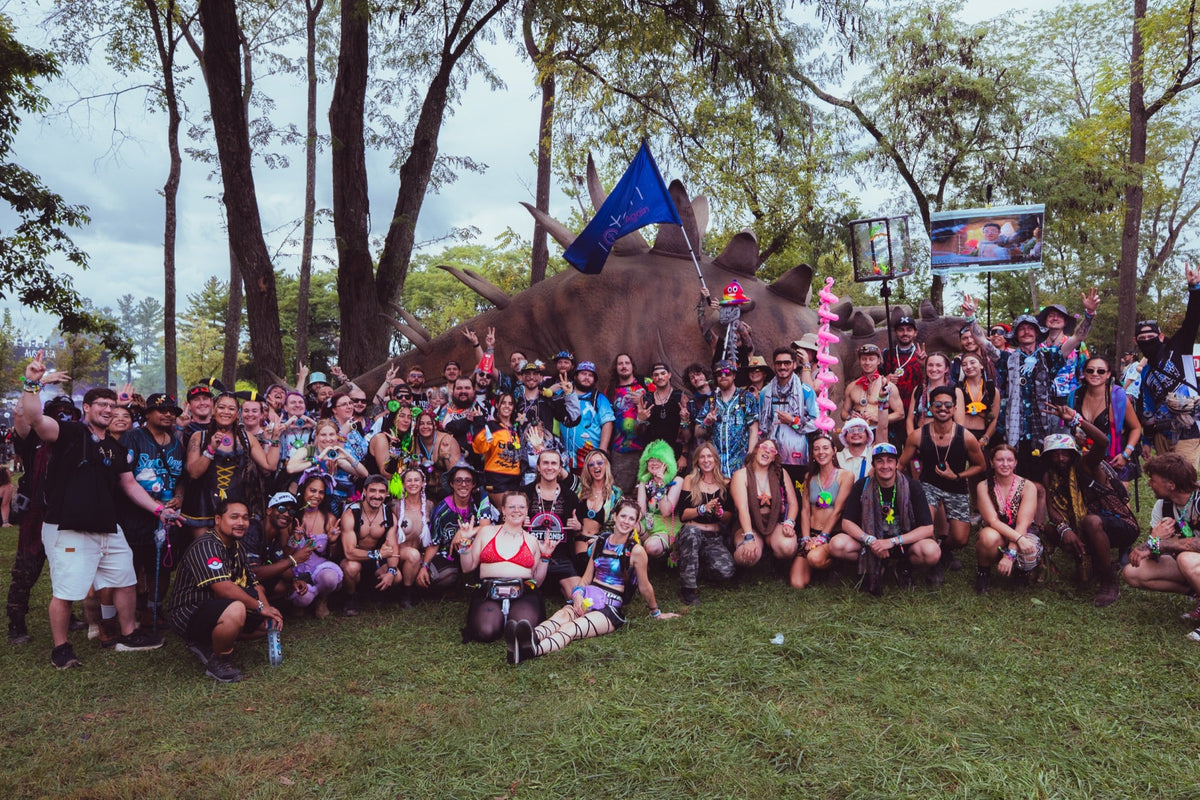 Large crowd of Totem owners in colorful festival outfits posing for a group photo in front of a giant stegosaurus statue at Lost Lands, with a blue Totem flag raised above them
