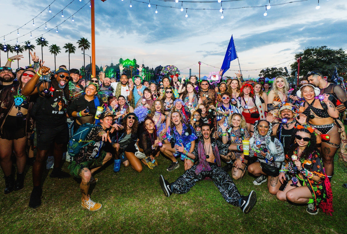 Large group of festival-goers in colorful outfits posing on the grass at a daytime Totem meetup, with palm trees and a blue Totem flag behind them