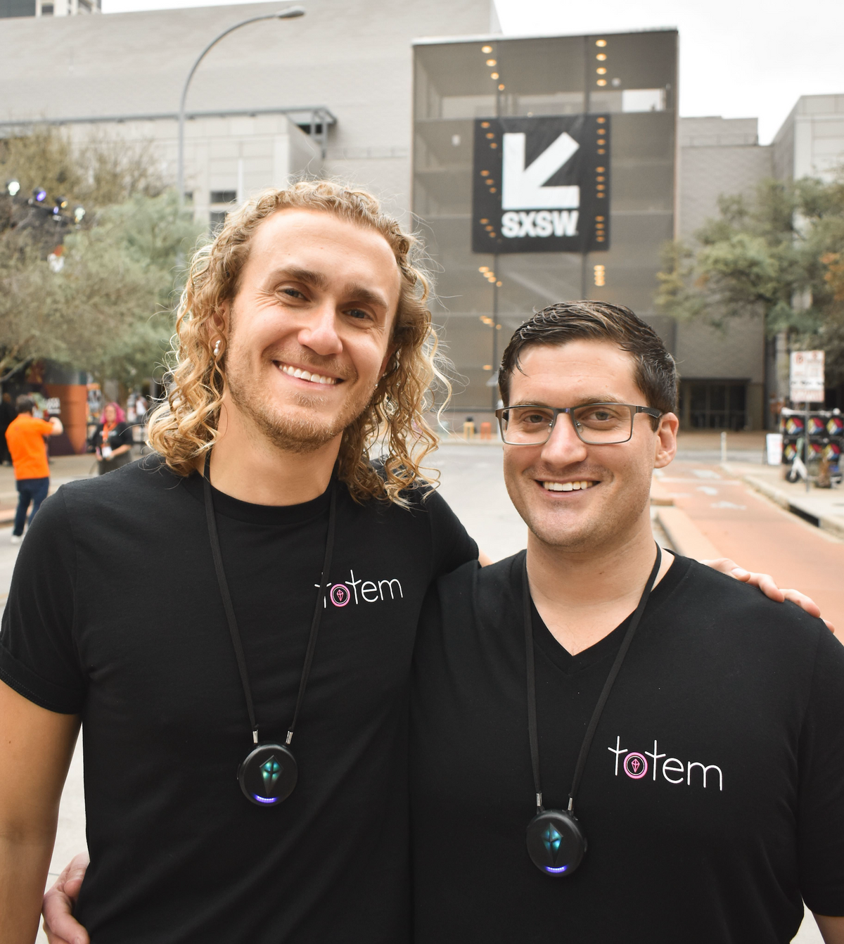Two smiling Totem team members in black Totem T-shirts, each wearing a glowing Totem Compass necklace, outside the SXSW venue in Austin