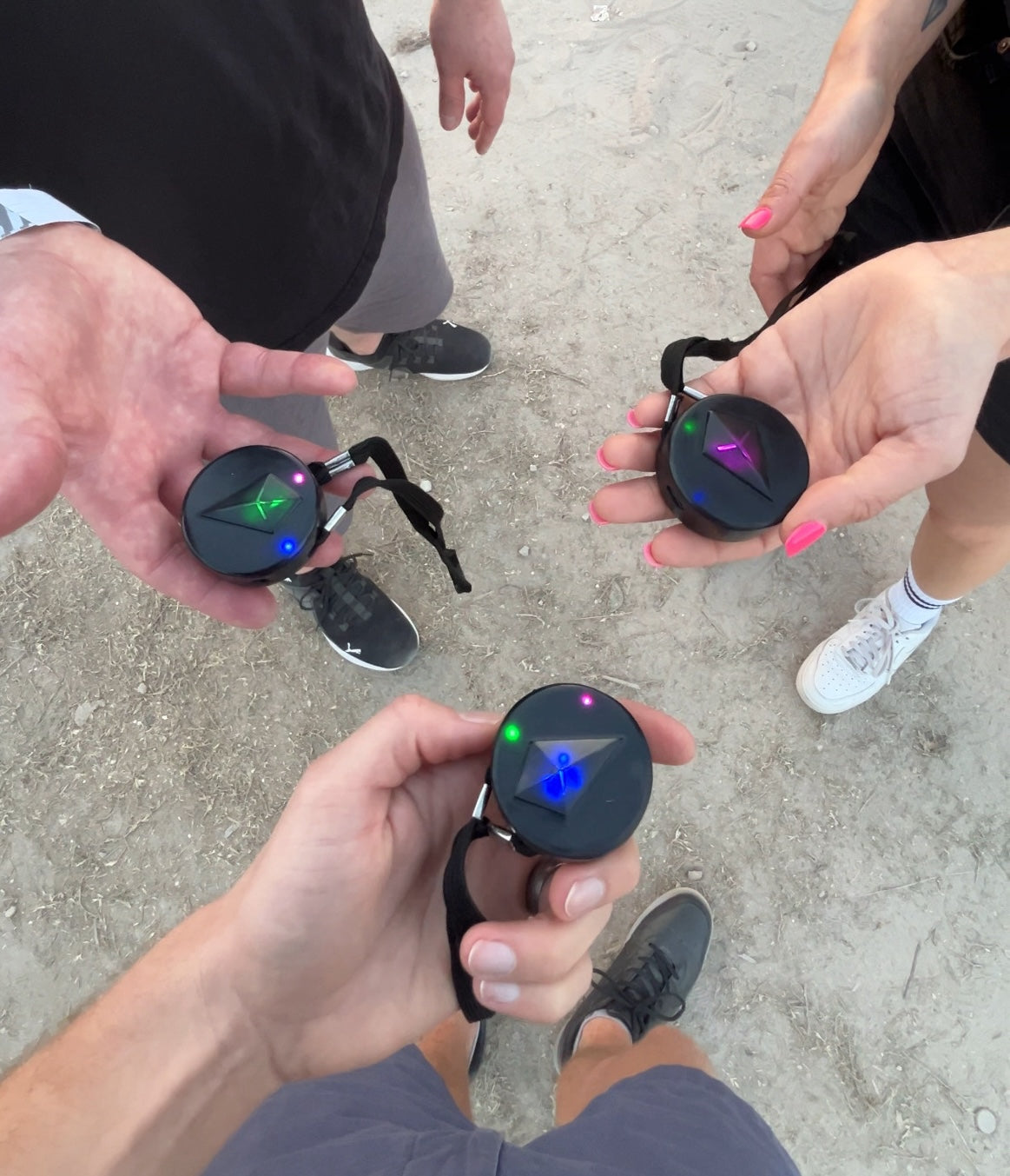 Three people's hands holding Totem Compasses toward each other over sandy ground, each Compass showing colored Bond lights