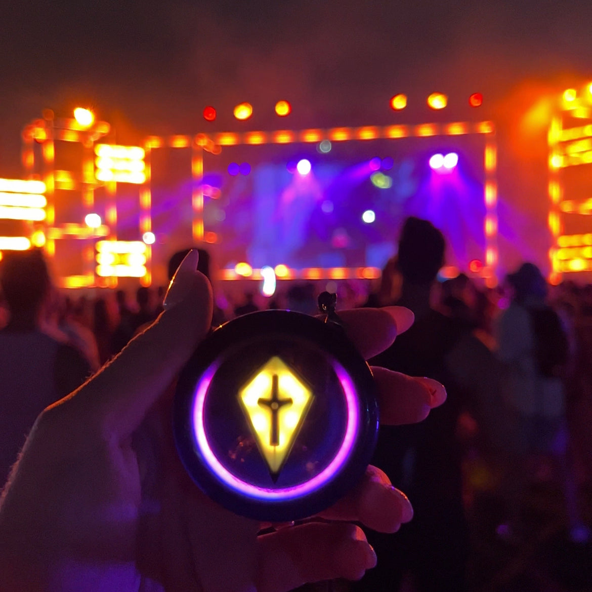 Hand holding up a Totem Compass with a yellow Touch Crystal and purple ring in front of a brightly lit festival stage at night