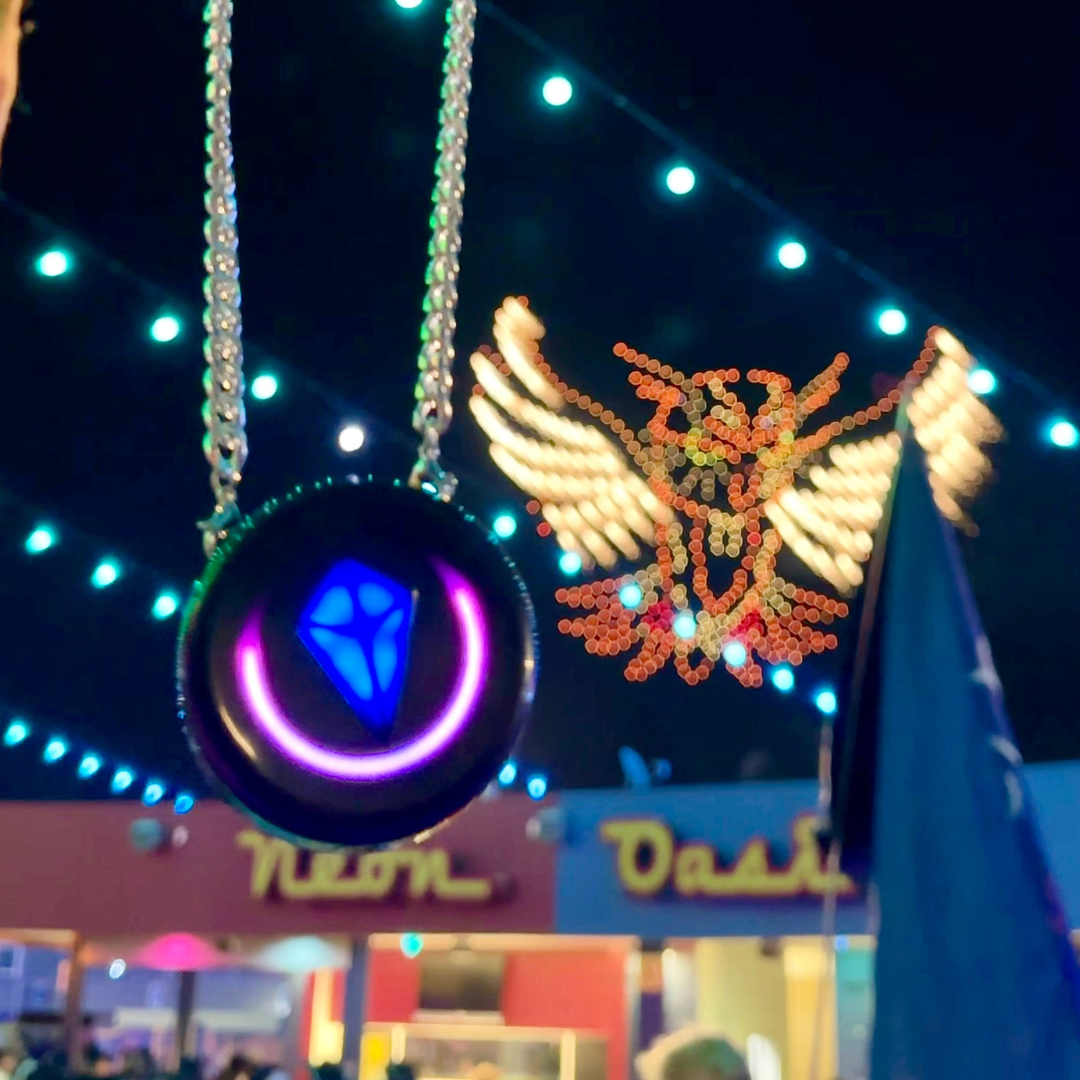 Totem Compass with a blue crystal and pink ring hanging from a silver chain at night, in front of a lit-up owl sculpture and a "Neon Oasis" sign
