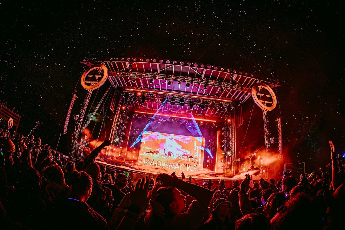 Festival crowd at night facing a stage lit orange and red, with Dirtybird Campout logos on either side and "Badsnacks" on the screen
