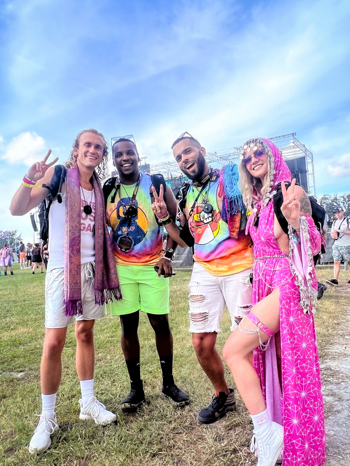 Four friends in tie-dye and festival outfits flash peace signs in front of a stage, several wearing Totem Compasses on lanyards
