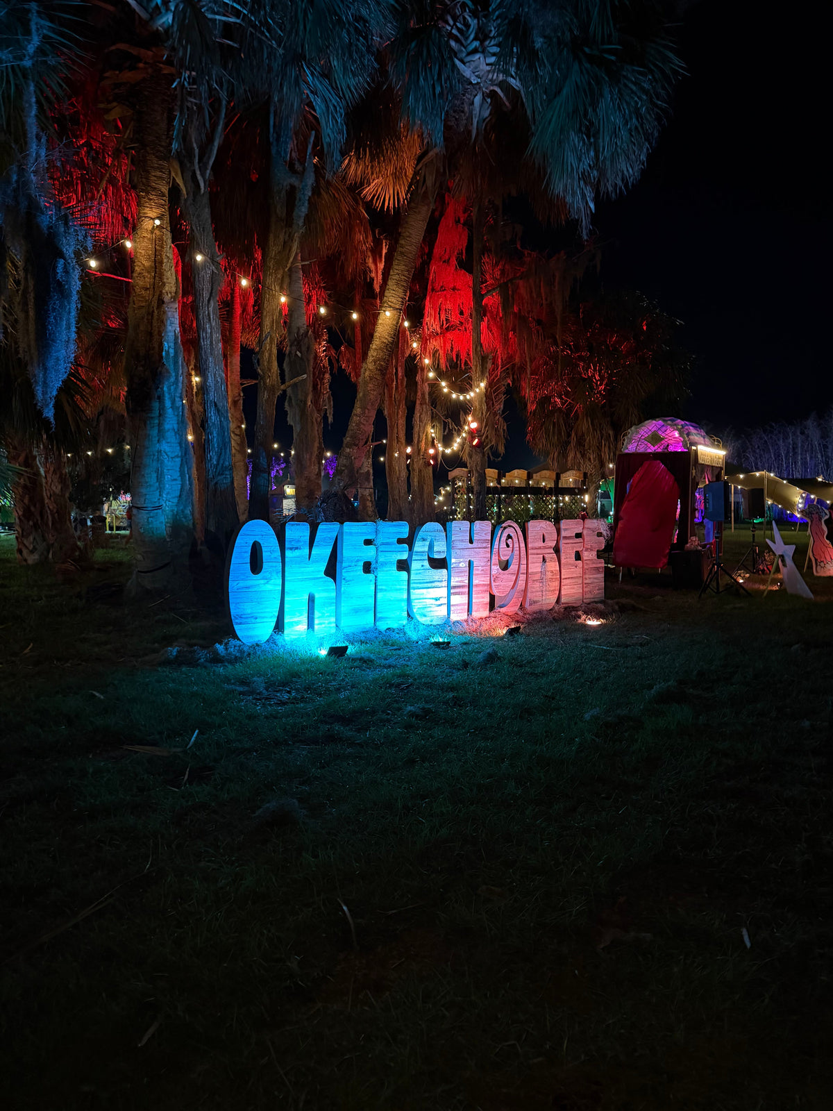 Illuminated OKEECHOBEE letter sign glowing blue and pink beneath palm trees strung with lights at night